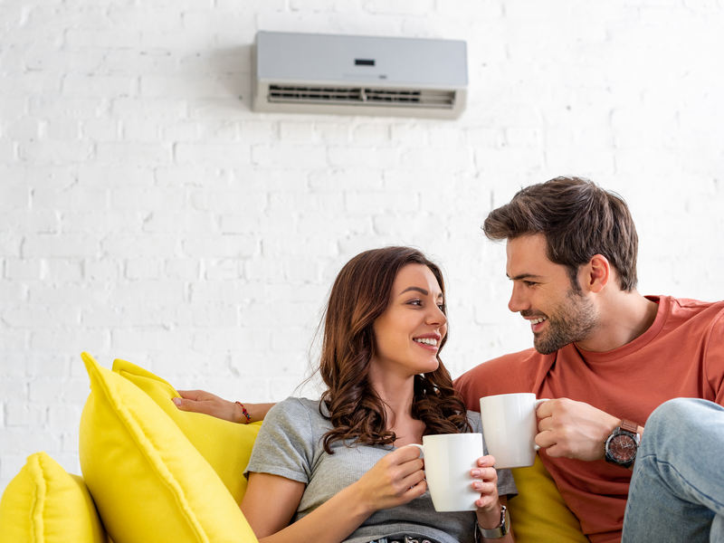 Happy,Couple,With,Cups,Sitting,On,Sofa,Under,Air,Conditioner